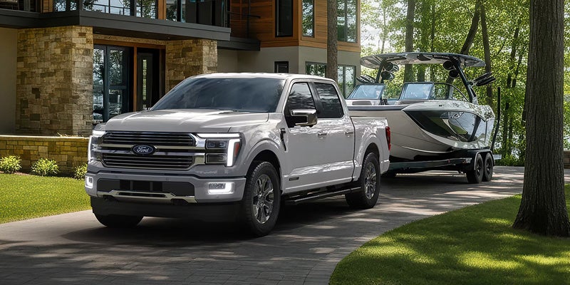 A Ford F-150 towing a boat on a trailer, parked near a lake with trees in the background.